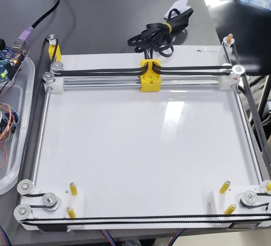 The first, smaller version of the CoreXY gantry on a white base, with the yellow carriage and belts around the edges